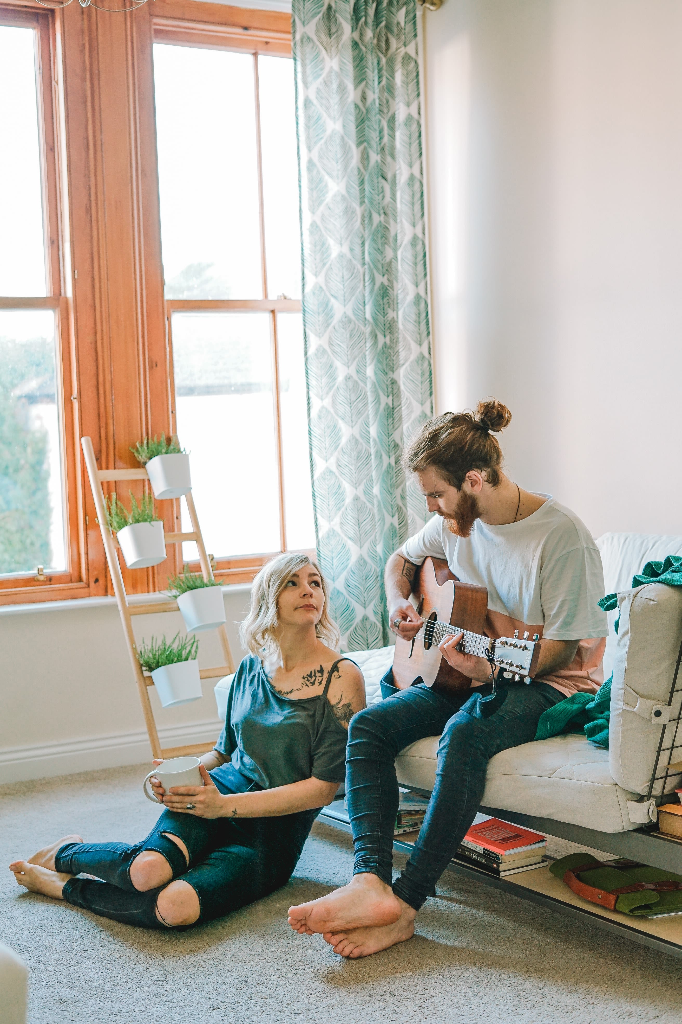 man sitting on sofa playing guitar looking at girl sitting on the ground near window inside room